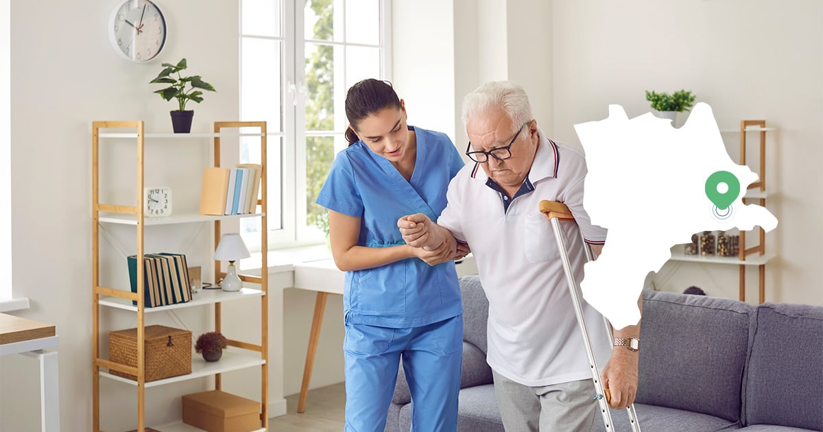 An elderly man with a crutch is helped to walk by a carer at home