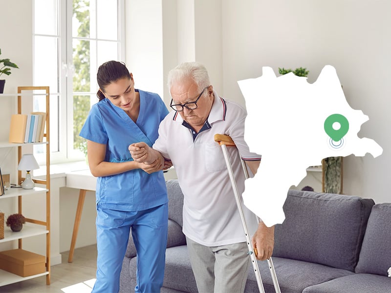 An elderly man with a crutch is helped to walk by a carer at home