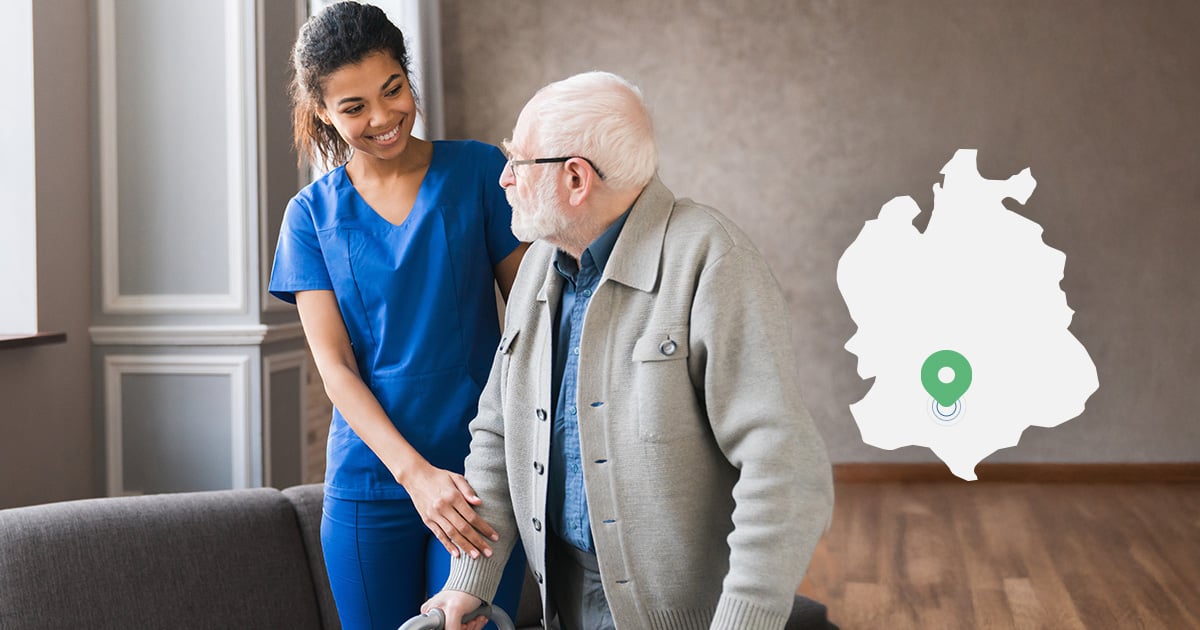 An elderly man is helped to walk at home by a carer.
