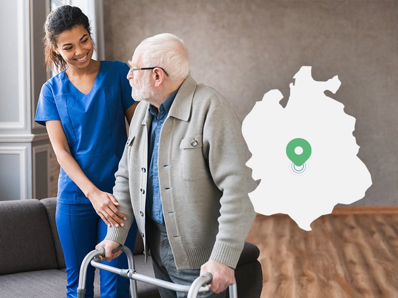 An elderly man is helped to walk at home by a carer.