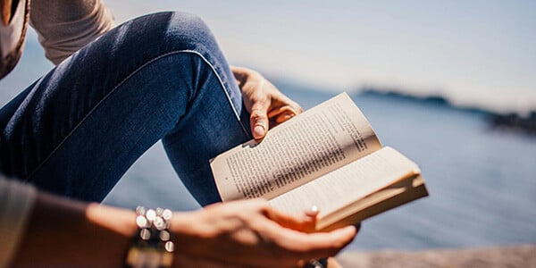 Woman sitting by the water with a book in her hand, practicing self-care as a family caregiver