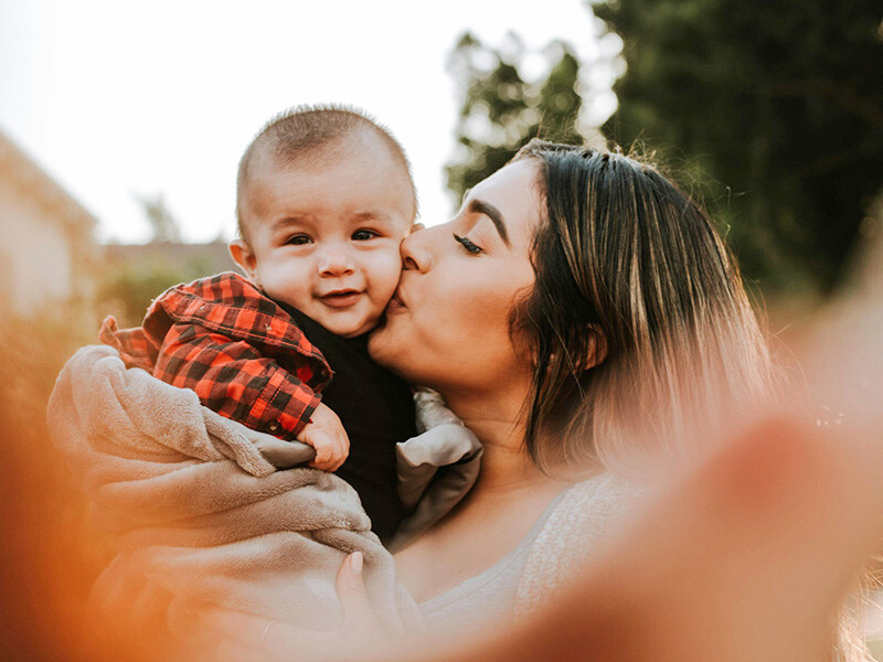 A young woman lovingly kisses a baby on the cheek, both surrounded by warm sunlight, a moment of familial tenderness and bonding.