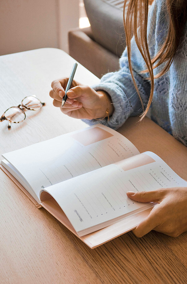 A woman in a blue sweater is writing in an open planner, next to her are a pair of glasses, a symbol of trust-based working hours.