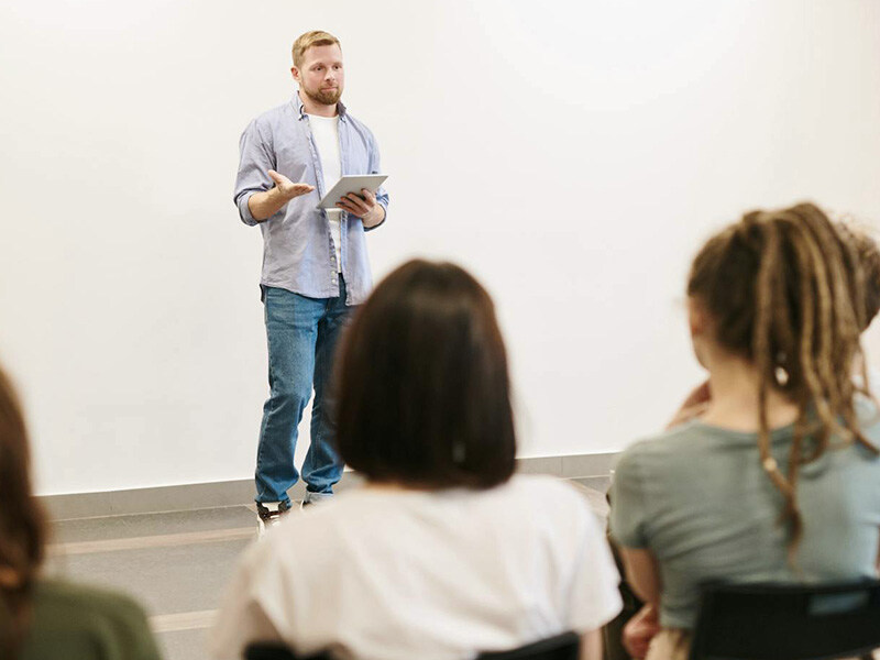 A man with a tablet in his hand presents to a group of seated listeners, an example of professional training and employee development.