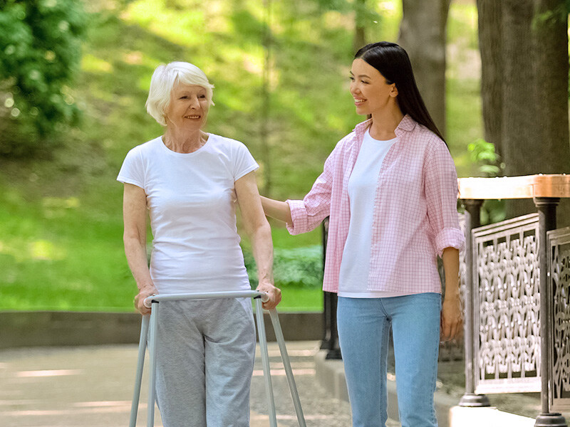 A young woman accompanies an elderly lady with a walking frame on a walk in a sunny park.