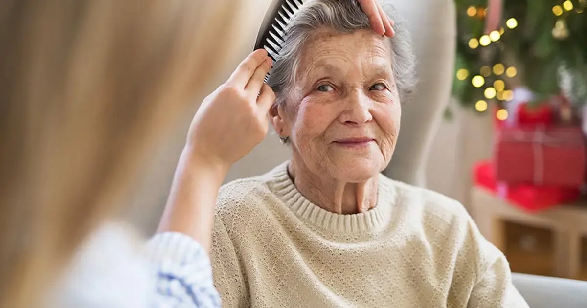 A nursing assistant lovingly combs the hair of a smiling elderly woman sitting in a cozy room decorated for Christmas.