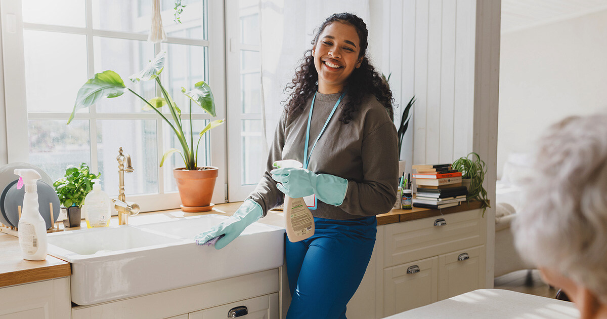 A smiling housekeeper wearing protective gloves cleans the kitchen while an elderly person sits in the foreground.