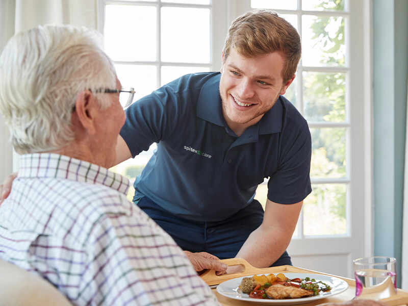 A care professional in a blue shirt smiles at an elderly gentleman as he serves him a meal.