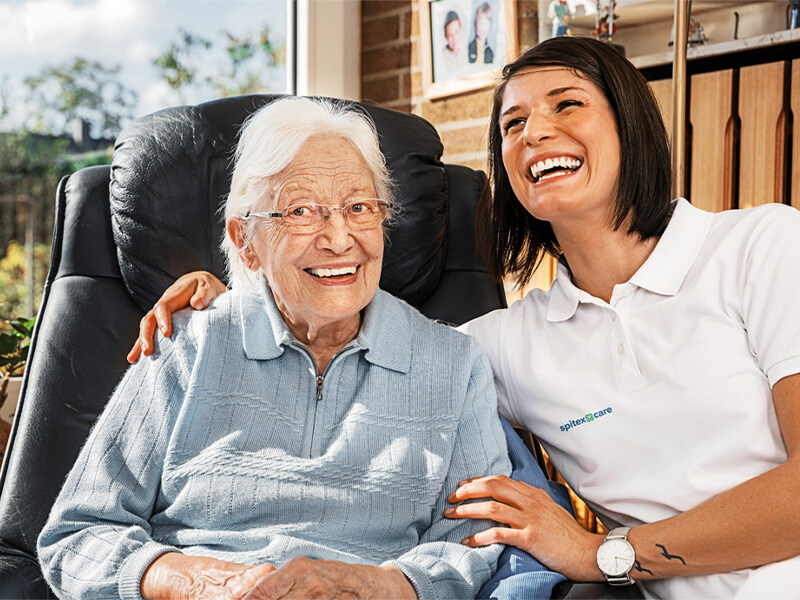 Healthcare specialist laughs with an elderly lady sitting in a black leather armchair in a bright, homely room