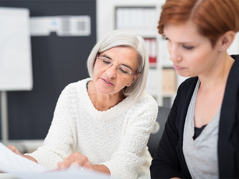 Two women are sitting together at the table, looking intently at a document that the older woman is holding in her hand.