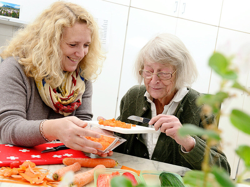 A housekeeping employee with curly blonde hair helps an elderly lady with glasses to cut vegetables in a kitchen