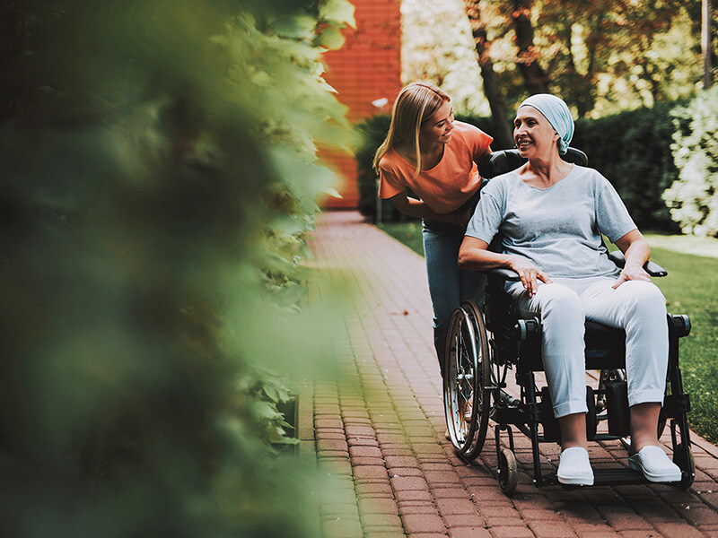 un woman pushes smiling elderly lady in a headscarf in a wheelchair through a green park, a picture of support in oncology care.