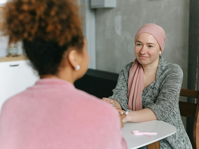 Two women in a reassuring conversation, one with a headscarf sits opposite smiling confidently, a moment of caring in oncology care.