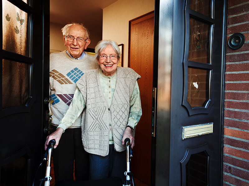 An elderly couple stand smiling in the doorway of their house, both using walking aids. They are standing in front of a dark front door.