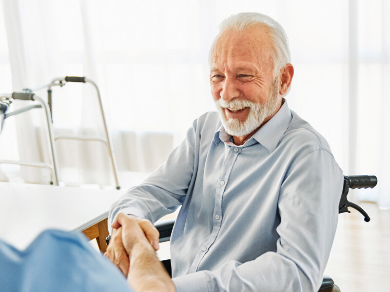 Smiling elderly man in a wheelchair shakes the hand of a care manager in a bright apartment, a walking frame in the background.