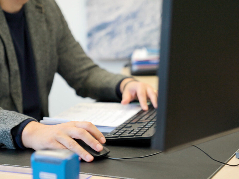 Nursing manager at a desk, operating a keyboard and a computer mouse, office and monitor can be seen blurred in the background.