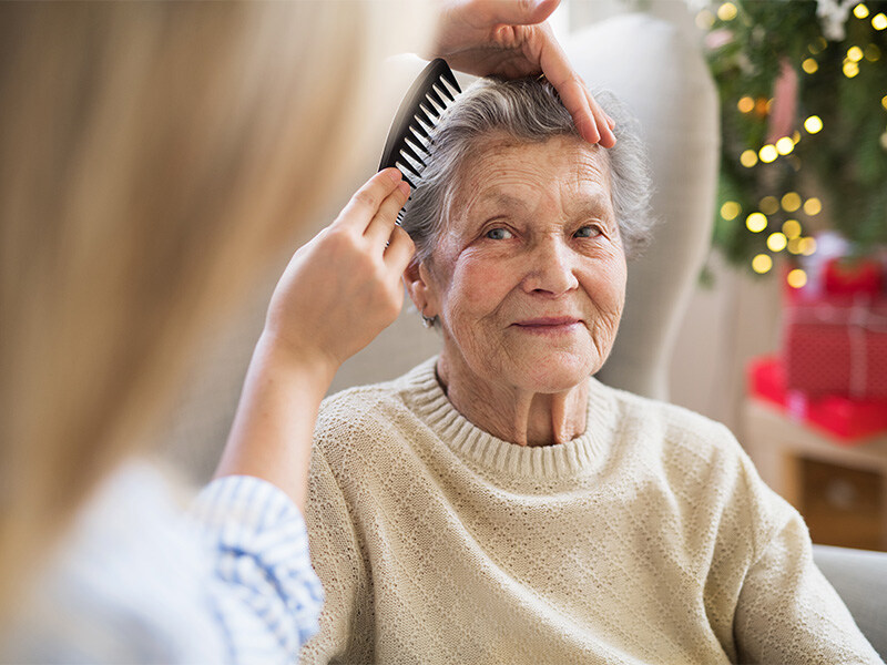 A care assistant lovingly combs the hair of a smiling elderly woman sitting in a cozy room with Christmas decorations.