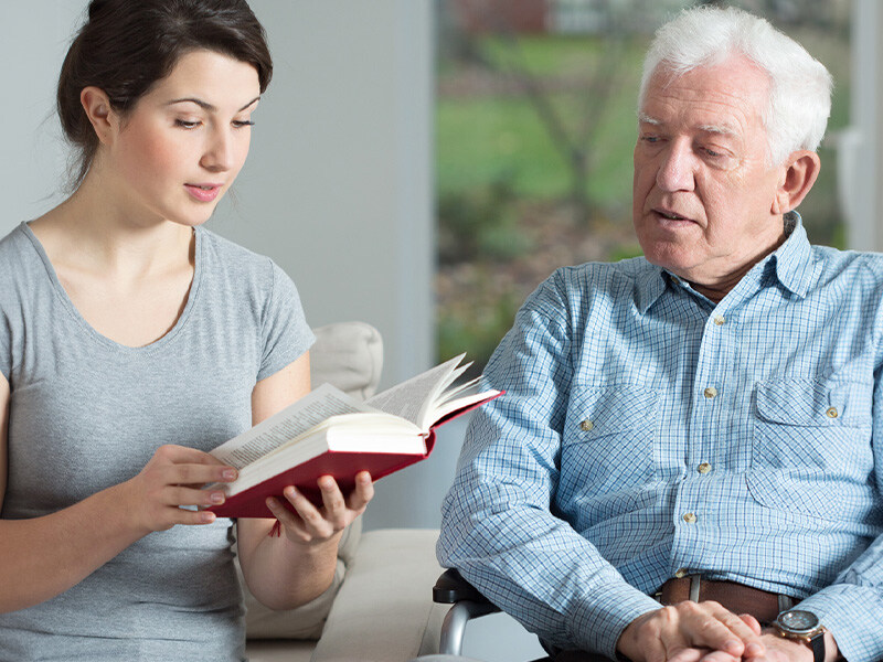 A young woman reads a book to an elderly man sitting in a wheelchair as an example of caring for parents at home.