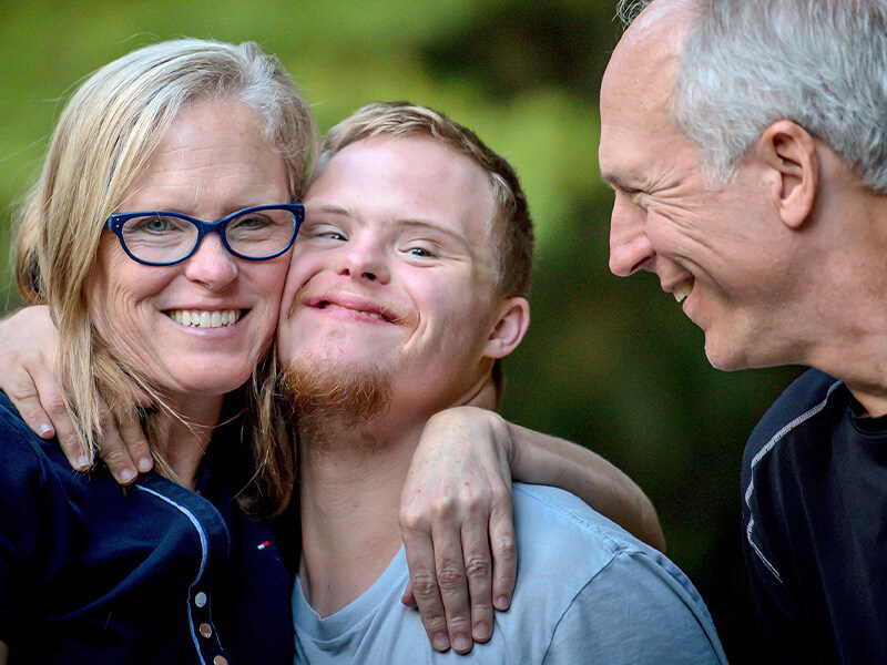 Smiling caring relatives lovingly hug their adult son with Down syndrome outdoors.