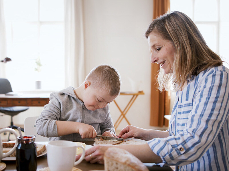 Mother and child with Down syndrome eat breakfast together, mother helps child to butter bread