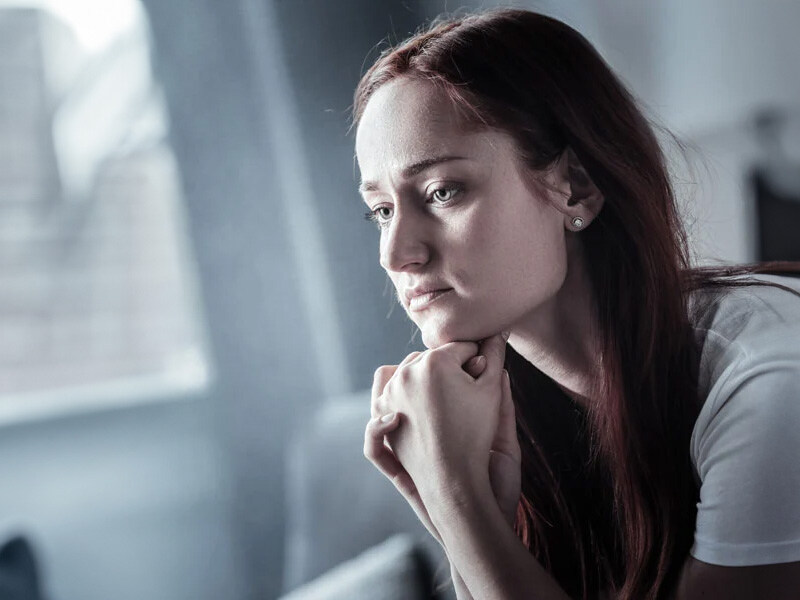 A thoughtful young woman with red hair and a white top puts her hand to her chin and looks worried