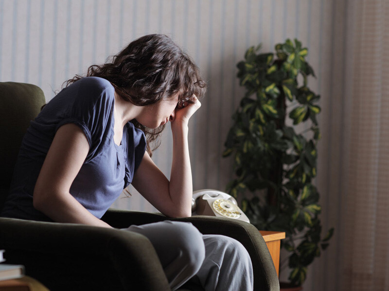 Woman in psychiatric care sits thoughtfully on a chair next to a retro telephone and a houseplant.