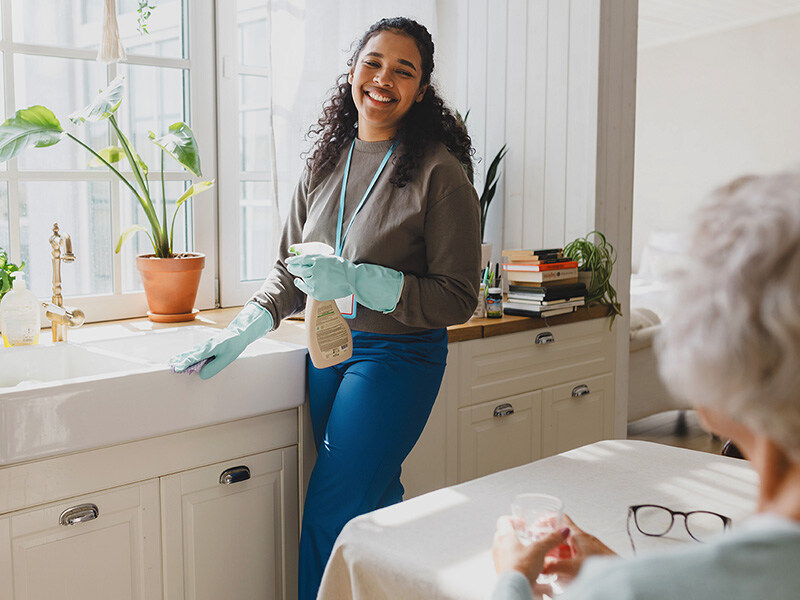 Smiling domestic help with protective gloves cleans kitchen while an elderly person sits in the foreground.