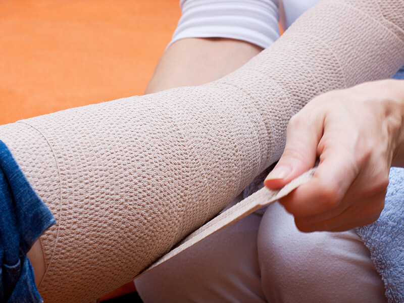 A close-up of a person applying an elastic medical bandage to a leg. The person is seated and wearing white clothing.