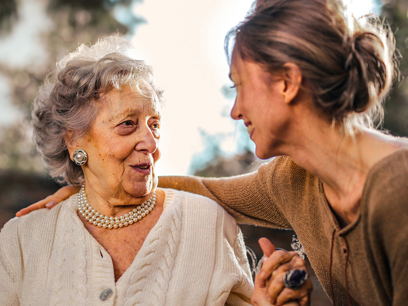 An elderly lady with a pearl necklace smiles gratefully as she is lovingly embraced by a younger woman, presumably a carer.