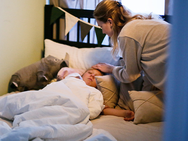 A young woman bends over a sleeping child in bed, a picture of domestic care by relatives.