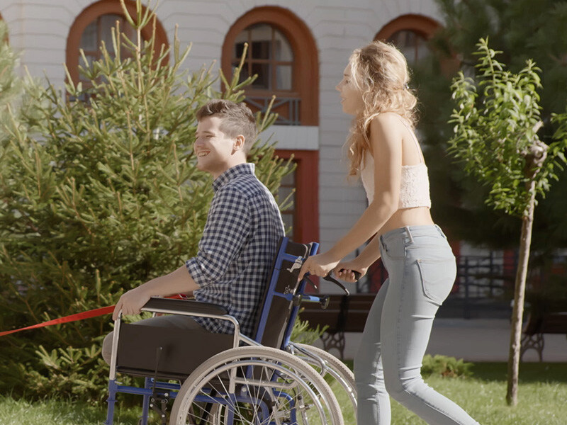 Young woman pushes young man in wheelchair, man holds red leash, both could be a couple