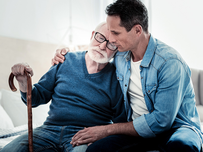 An elderly gentleman with glasses and a blue sweater leans against his son, who is hugging him, in a moment of care by caring relatives.